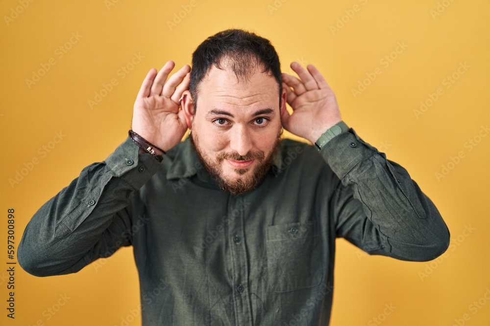 Plus size hispanic man with beard standing over yellow background trying to hear both hands on ear gesture, curious for gossip. hearing problem, deaf Plus size hispanic man with beard standing over yellow background trying to hear both hands on ear gesture, curious for gossip. hearing problem, deaf