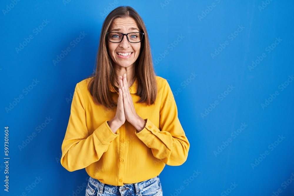 Young woman wearing glasses standing over blue background praying with hands together asking for forgiveness smiling confident. Young woman wearing glasses standing over blue background praying with hands together asking for forgiveness smiling confident.