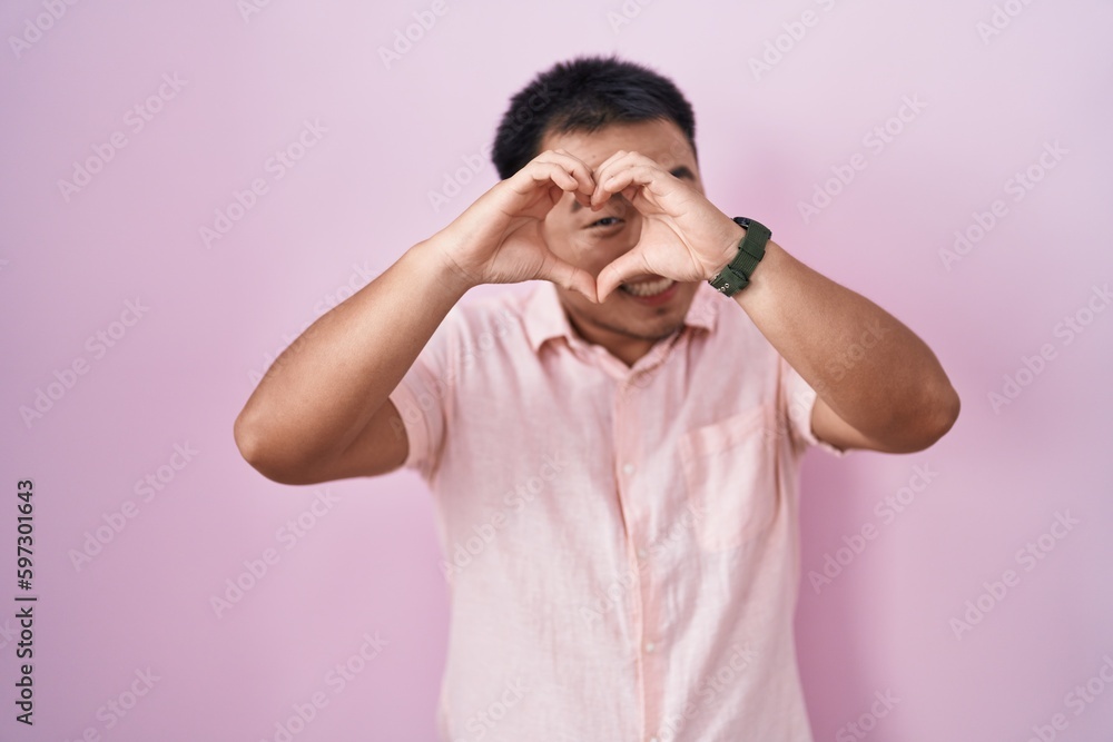 Chinese young man standing over pink background doing heart shape with hand and fingers smiling looking through sign Chinese young man standing over pink background doing heart shape with hand and fingers smiling looking through sign
