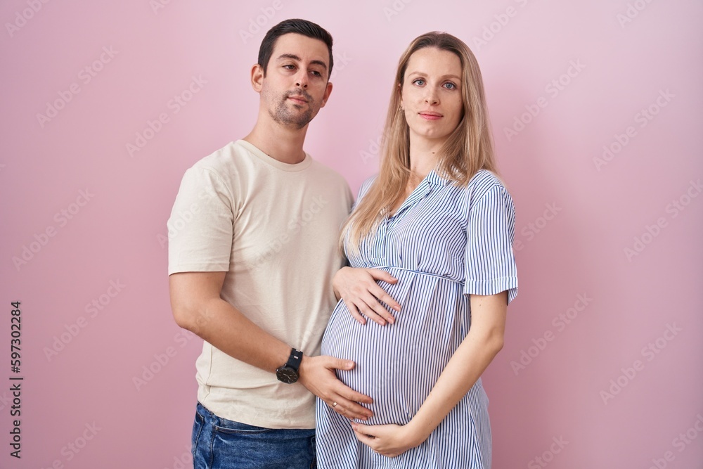 Young couple expecting a baby standing over pink background relaxed with serious expression on face. simple and natural looking at the camera. Young couple expecting a baby standing over pink background relaxed with serious expression on face. simple and natural looking at the camera.