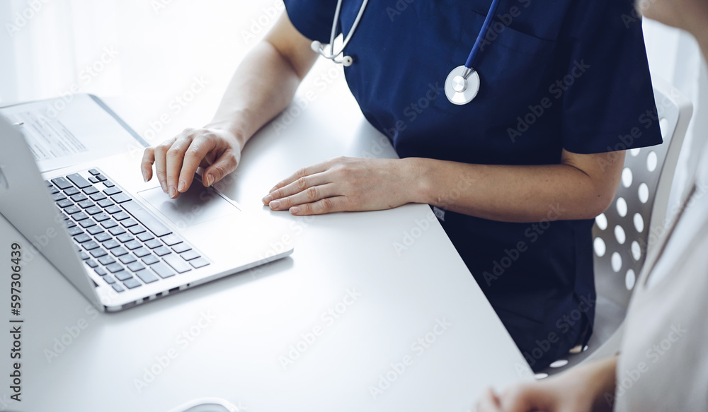 Doctor and patient sitting at the desk in clinic office. The focus is on unknown female physician's hands using a laptop computer, close up. Medicine concept Doctor and patient sitting at the desk in clinic office. The focus is on unknown female physician's hands using a laptop computer, close up. Medicine concept