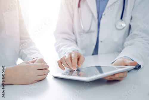 Doctor and patient sitting near each other at the desk in clinic. The focus is on female physician s hands pointing into tablet computer touchpad  close up. Medicine concept