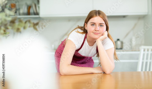 in modern kitchen interior  smiling young girl in red apron with polka dots props her chin with her fist and leans on kitchen table. Housework  waiting  planning