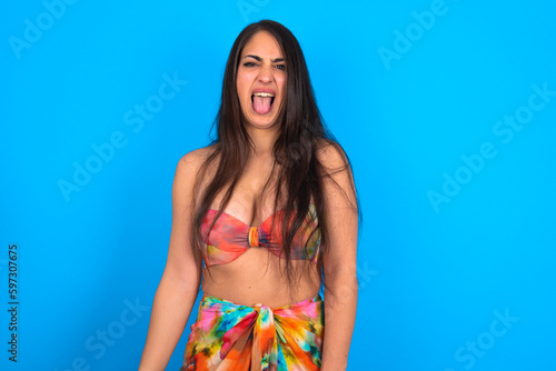 beautiful brunette woman wearing bikini over blue background sticking tongue out happy with funny expression. Emotion concept.