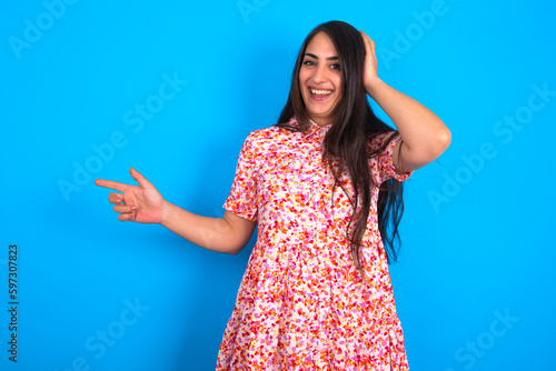 Surprised beautiful brunette woman wearing floral dress over blue background pointing at empty space holding hand on head
