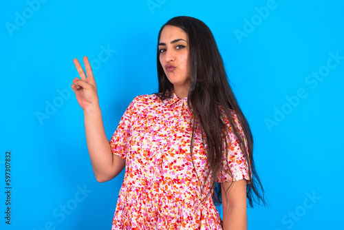 beautiful brunette woman wearing floral dress over blue background makes peace gesture keeps lips folded shows v sign. Body language concept