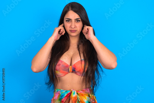 beautiful brunette woman wearing swimwear over blue background covering ears with fingers with annoyed expression for the noise of loud music. Deaf concept.