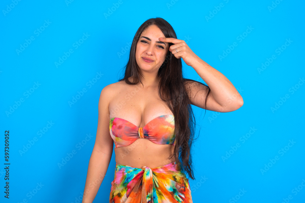 beautiful brunette woman wearing swimwear over blue background pointing unhappy at pimple on forehead, blackhead  infection. Skincare concept.