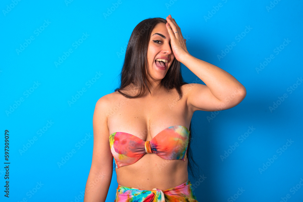 Obraz premium beautiful brunette woman wearing swimwear over blue background surprised with hand on head for mistake, remember error. Forgot, bad memory concept.