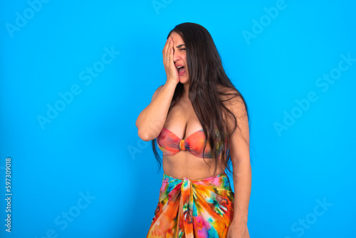 beautiful brunette woman wearing swimwear over blue background Yawning tired covering half face, eye and mouth with hand. Face hurts in pain.
