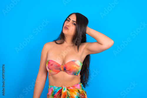 beautiful brunette woman wearing swimwear over blue background confuse and wonder about question. Uncertain with doubt, thinking with hand on head. Pensive concept.