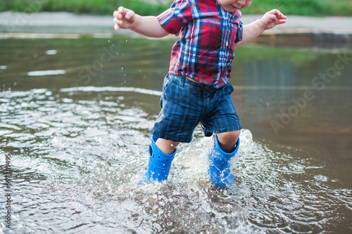 Happy toddler boy running through puddles in rubber boots. Child splashes after rain outside..