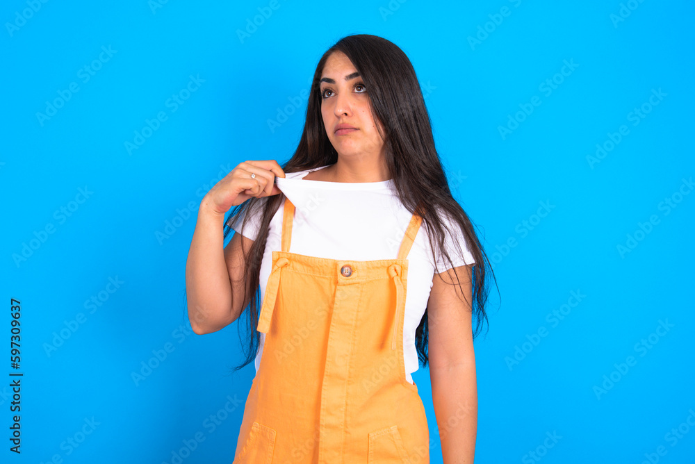 brunette woman wearing orange overalls over blue studio background stressed, anxious, tired and frustrated, pulling shirt neck, looking frustrated with problem brunette woman wearing orange overalls over blue studio background stressed, anxious, tired and frustrated, pulling shirt neck, looking frustrated with problem