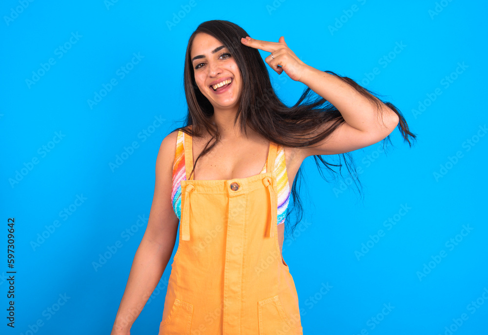 brunette woman wearing orange overalls over blue studio background foolishness around shoots in temple with fingers makes suicide gesture. Funny model makes finger gun pistol