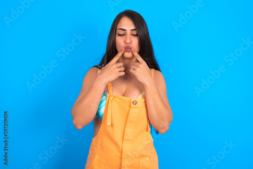 Canvas Print brunette woman wearing orange overalls over blue studio background crosses eyes