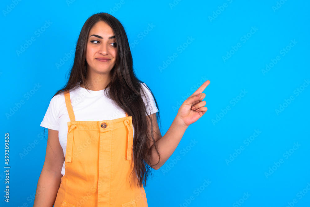 brunette woman wearing orange overalls over blue studio background points at copy space and advertises something, advices best price. brunette woman wearing orange overalls over blue studio background points at copy space and advertises something, advices best price.