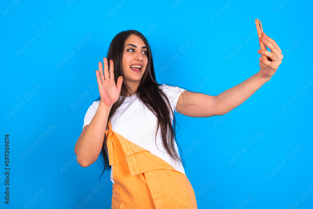 brunette woman wearing orange overalls over blue studio background holds modern mobile phone and makes video call waves palm in hello gesture. People modern technology concept
