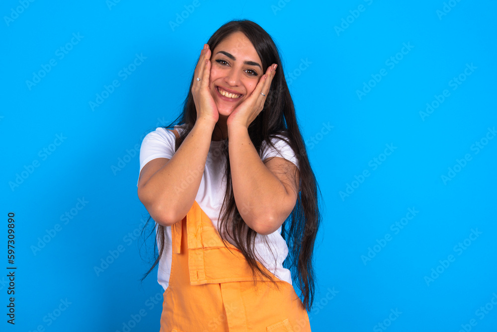 Happy brunette woman wearing orange overalls over blue studio background touches both cheeks gently, has tender smile, shows white teeth, gazes positively straightly at camera, Happy brunette woman wearing orange overalls over blue studio background touches both cheeks gently, has tender smile, shows white teeth, gazes positively straightly at camera,