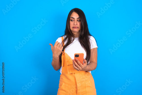 brunette woman wearing orange overalls over blue studio background points thumb away and shows blank space aside, holds mobile phone for sending text messages.