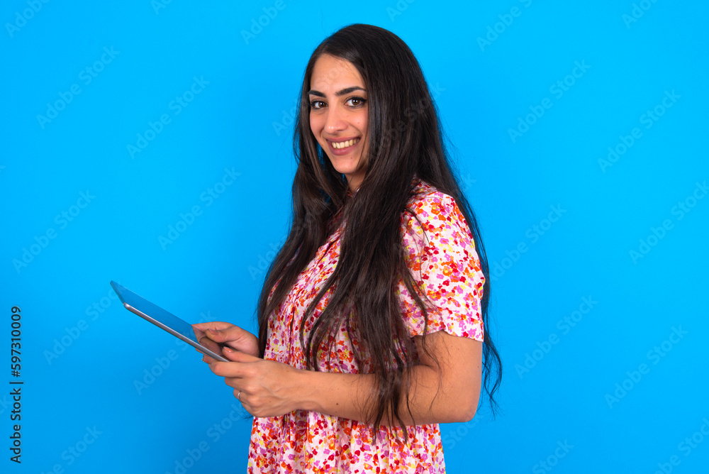 Photo of optimistic beautiful brunette woman wearing floral dress over blue background hold tablet Photo of optimistic beautiful brunette woman wearing floral dress over blue background hold tablet