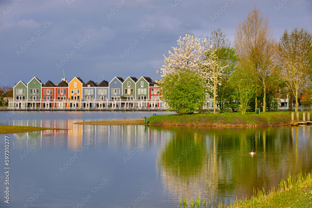 Houten, The Netherlands - April 25 2023. Trees and colourful wooden lakeside houses reflected in the water of lake De Rietplas. Houten, The Netherlands - April 25 2023. Trees and colourful wooden lakeside houses reflected in the water of lake De Rietplas.