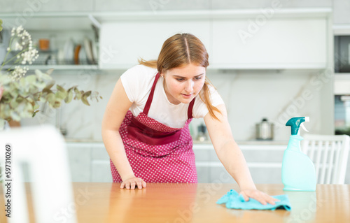 Smiling teenage girl in red retro polka dot apron using spray and wiping dust on countertop with microfiber rag while cleaning cozy cuisine