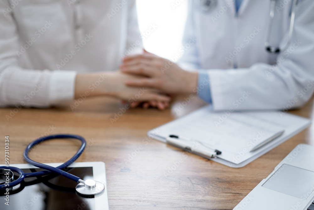 Stethoscope and laptop computer are lying on the wooden table while doctor's hands reassuring a patient at the background. Medicine concept Stethoscope and laptop computer are lying on the wooden table while doctor's hands reassuring a patient at the background. Medicine concept