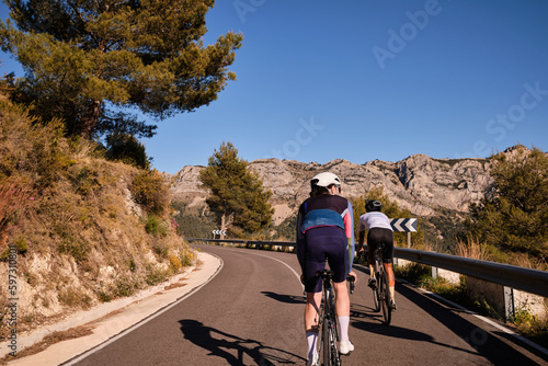 Two cyclists in full cycling gear and helmets ride their road bikes on mountain road at sunset Coll De Rates pass in Spain.Sportsmen training hard on bicycle outdoors.Sports motivation.