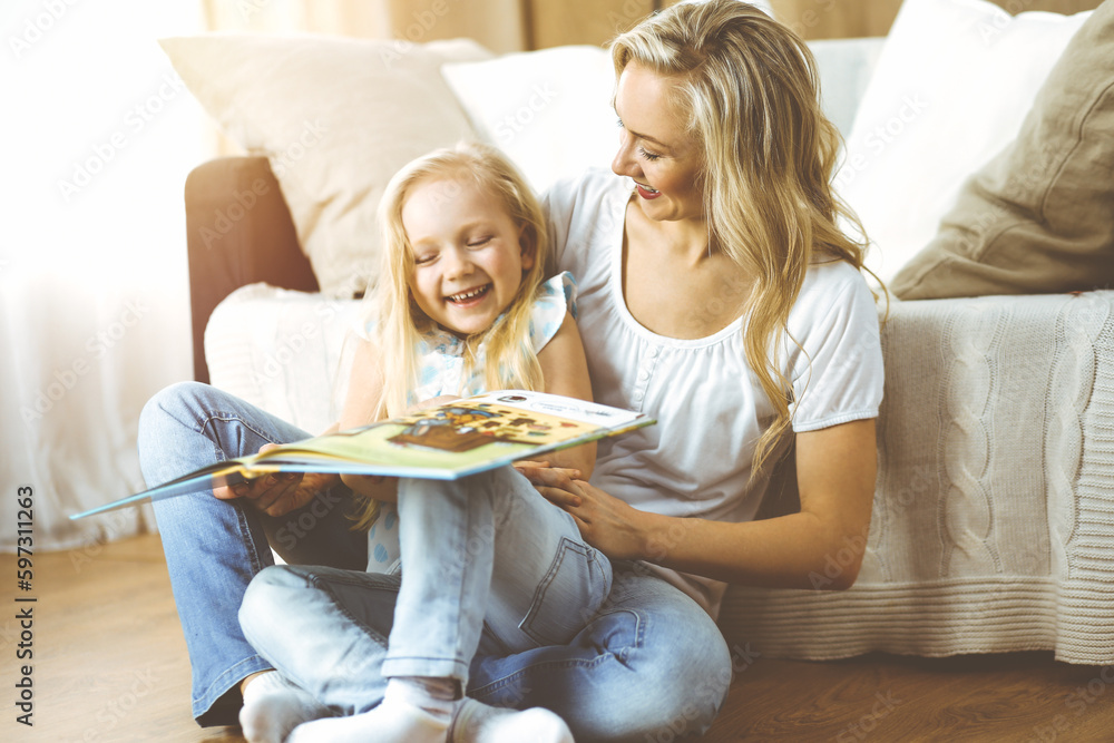 Happy family. Blonde young mother reading a book to her cute daughter while sitting at wooden floor in sunny room. Motherhood concept Happy family. Blonde young mother reading a book to her cute daughter while sitting at wooden floor in sunny room. Motherhood concept