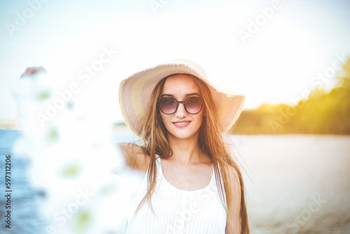 Happy smiling woman in free happiness bliss on ocean beach standing with hat, sunglasses, and white flowers. Portrait of a multicultural female model in white summer dress enjoying nature