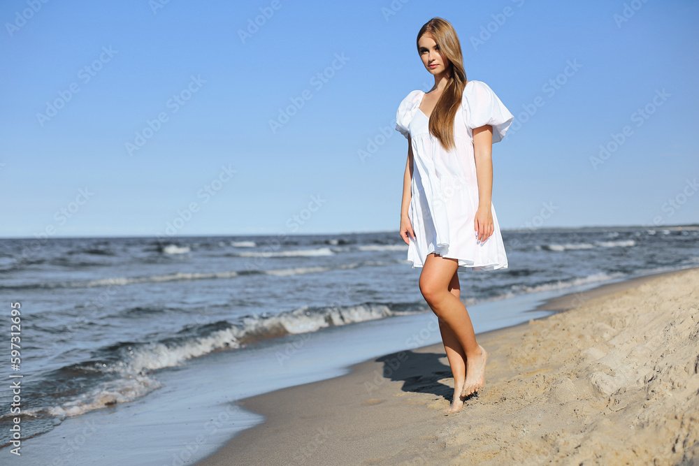 Happy smiling beautiful woman is walking on the ocean beach in a white summer dress