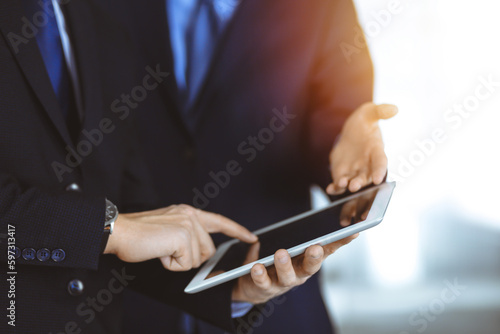 Business people use a tablet computer for discussion of their new project  standing in a sunny modern office. Unknown businessman or male entrepreneur with a colleague at workplace