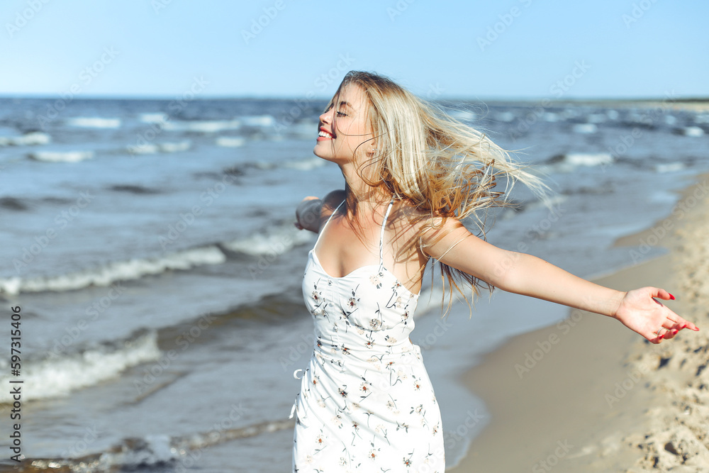 Happy blonde beautiful woman on the ocean beach standing in a white summer dress, raising hands Happy blonde beautiful woman on the ocean beach standing in a white summer dress, raising hands