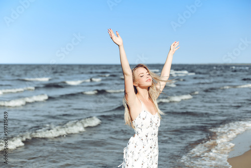 Happy blonde beautiful woman on the ocean beach standing in a white summer dress, raising hands
