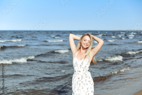 Fototapeta Naklejka Na Ścianę i Meble -  Happy blonde beautiful woman on the ocean beach standing in a white summer dress, raising hands