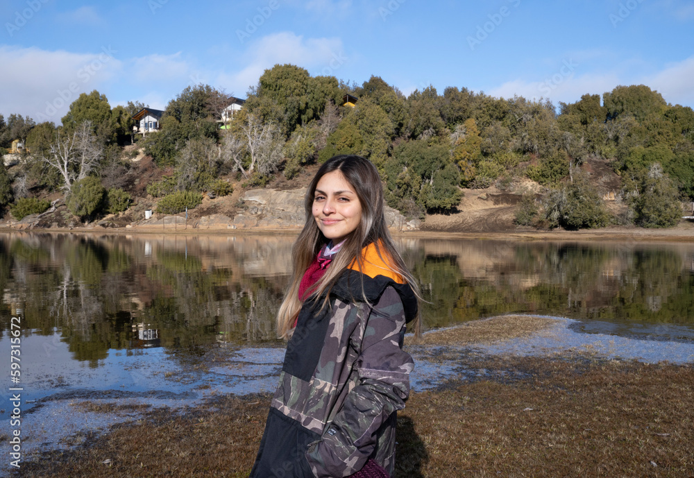 Portrait of a smiling young woman wearing a coat with the lake and forest in the background.
