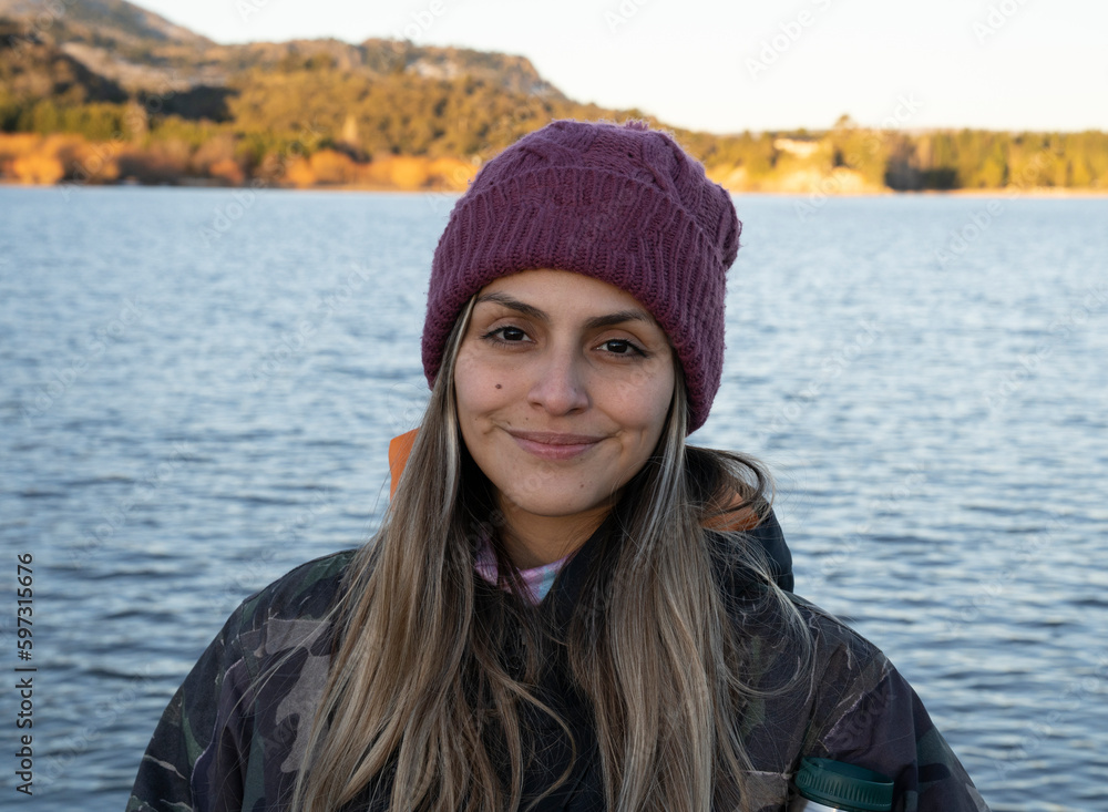 Portrait of a smiling young woman wearing a coat and purple hat with the lake and mountains in the background. Portrait of a smiling young woman wearing a coat and purple hat with the lake and mountains in the background.