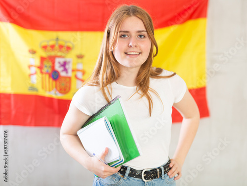 Happy smiling teen girl, student in white t-shirt and blue jeans holding spiral notepads under her arm against of national flag of Spain photo