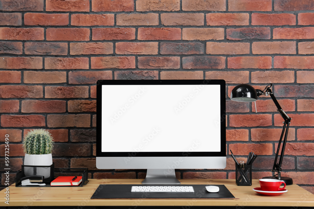 Fototapeta premium Stylish workplace with computer, lamp and stationery on wooden table near brick wall