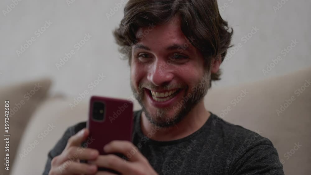 Excited young man jumps for joy as he receives good news on his smartphone while comfortably seated on his living room couch. Thrilled by this unexpected success Excited young man jumps for joy as he receives good news on his smartphone while comfortably seated on his living room couch. Thrilled by this unexpected success