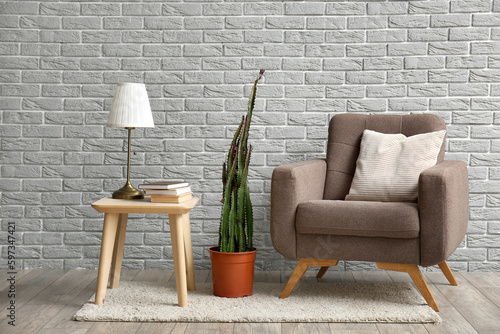 Cozy armchair, pot with cactus and lamp on table near grey brick wall