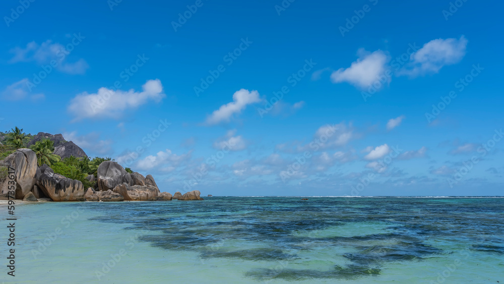 Picturesque granite boulders with smoothed outlines pile up at the edge of the ocean on the beach. Clear turquoise water. Green hill on the background of azure sky and clouds. Seychelles. La Digue.