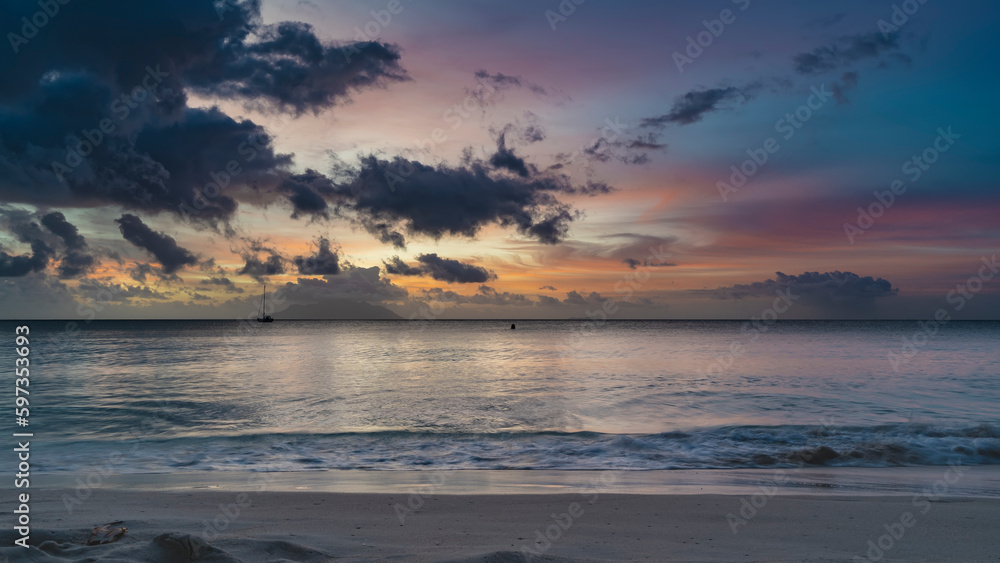 Colorful romantic sunset on a tropical beach. Waves are foaming on the sand. Purple clouds in the sky, illuminated with scarlet, pink, orange. Silhouettes of yachts and islands on the horizon.