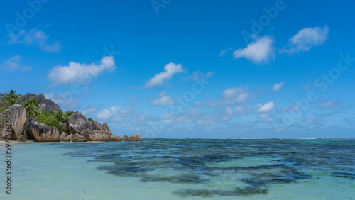 Picturesque granite boulders with smoothed outlines pile up at the edge of the ocean on the beach. Clear turquoise water. Green hill on the background of azure sky and clouds. Seychelles. La Digue.