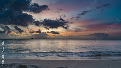 Colorful romantic sunset on a tropical beach. Waves are foaming on the sand. Purple clouds in the sky, illuminated with scarlet, pink, orange. Silhouettes of yachts and islands on the horizon.