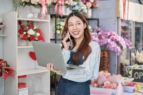 One young Asian female florist owner, working with laptop, selling floral arrange, talking on mobile phone in colorful flower shop store with a beautiful bunch of blossoms, and e-commerce business.