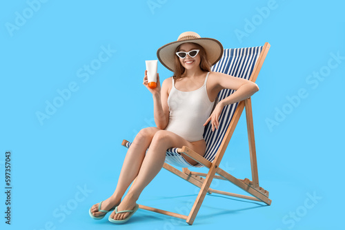 Beautiful young woman with sunscreen cream sitting on deck chair against blue background © Pixel-Shot