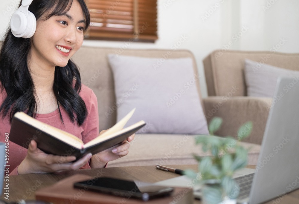 Student holding text book.Smiling asian girl wearing headphones using laptop, looking at screen, happy young female listening to music while working online. excited student learning language, Student holding text book.Smiling asian girl wearing headphones using laptop, looking at screen, happy young female listening to music while working online. excited student learning language,
