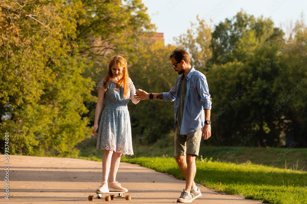 Young man embracing and helping girlfriend riding longboard on asphalt walkway between trees