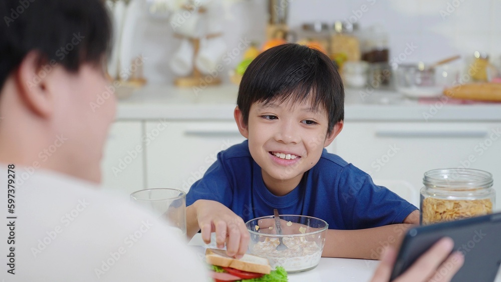 Little boy sitting at the table during breakfast at home in the kitchen waching and enjoy cartoon on tablet with father on weekend. Happy asian family Little boy sitting at the table during breakfast at home in the kitchen waching and enjoy cartoon on tablet with father on weekend. Happy asian family